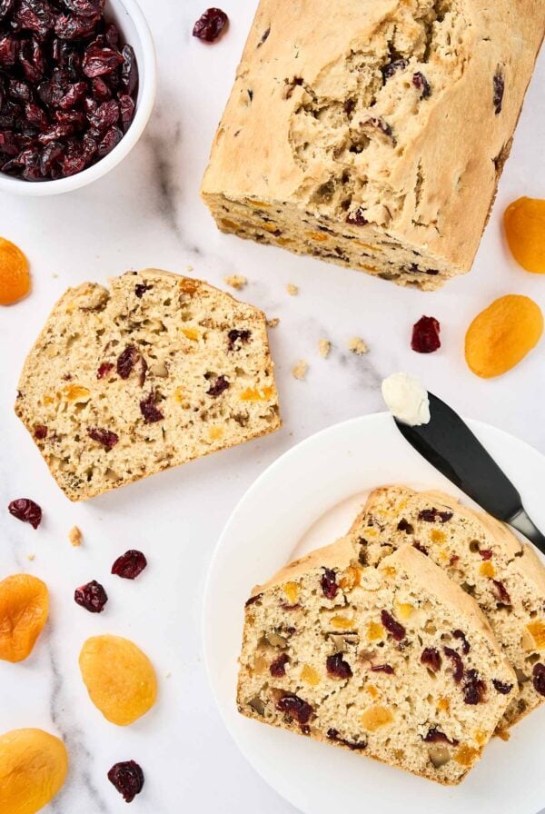 Sliced apricot bread with cranberries and walnuts on a white marble surface, surrounded by dried apricots, cranberries, and a buttered knife.