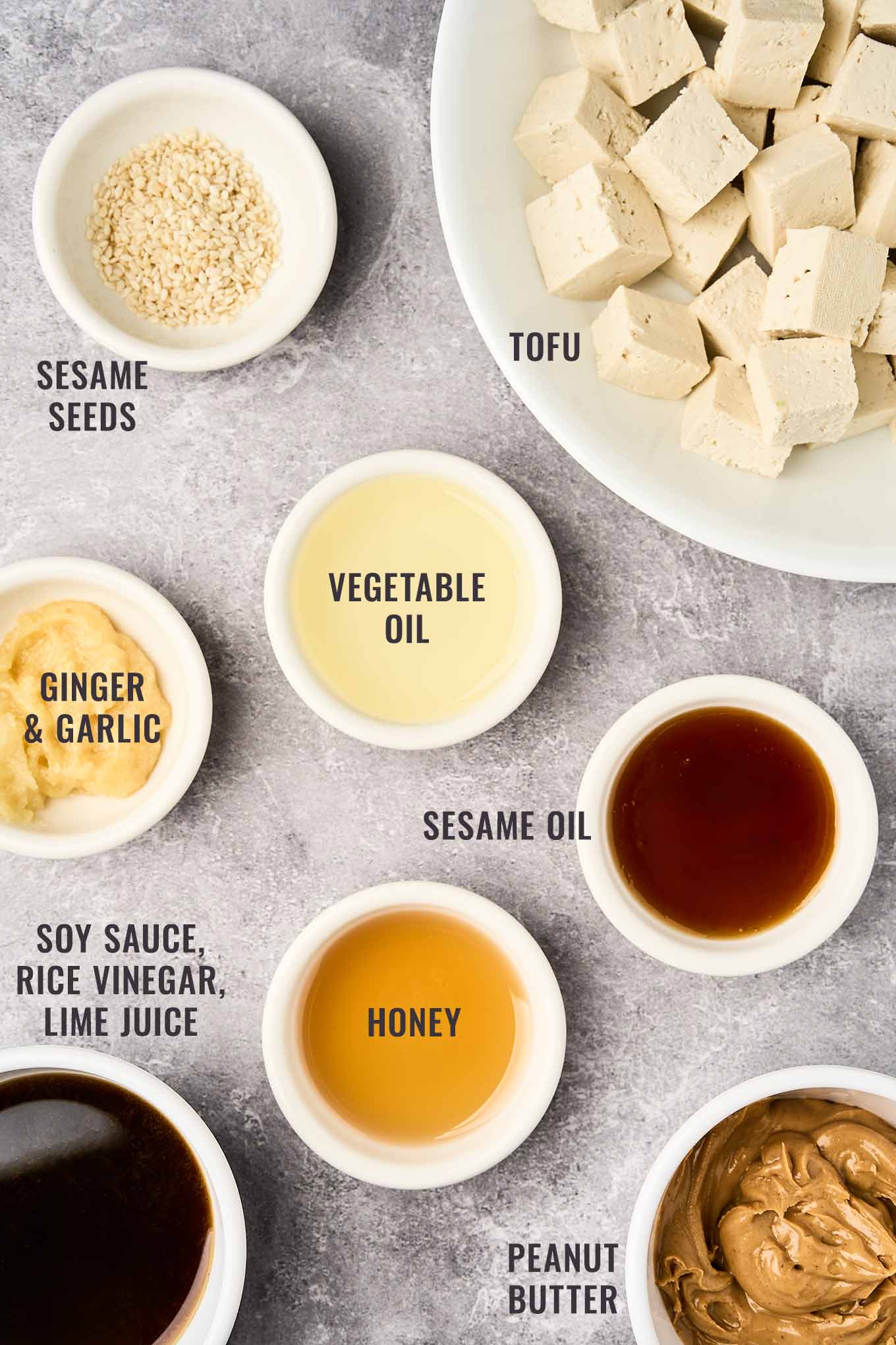 A labeled, overhead view of the ingredients for the peanut dressing and crispy tofu in Asian noodle salad: peanut butter, soy sauce, honey, lime juice, garlic, ginger, tofu, sesame seeds, and more.