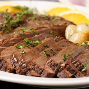 A close view of sliced braised brisket on a serving platter with pearl onions and a garnish of fresh thyme.