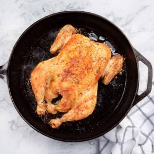 A golden brown, roasted chicken in a cast iron skillet on a marble surface.