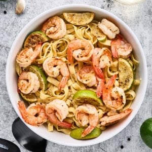 Garlic lime shrimp pasta in a white bowl, with sautéed shrimp, lime wedges, and fettuccine noodles in sharp focus.