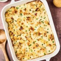 Golden-browned cottage cheese potato casserole in a white baking dish, topped with melted cheese and chopped chives, on a rustic surface with whole potatoes and a wooden spoon nearby.