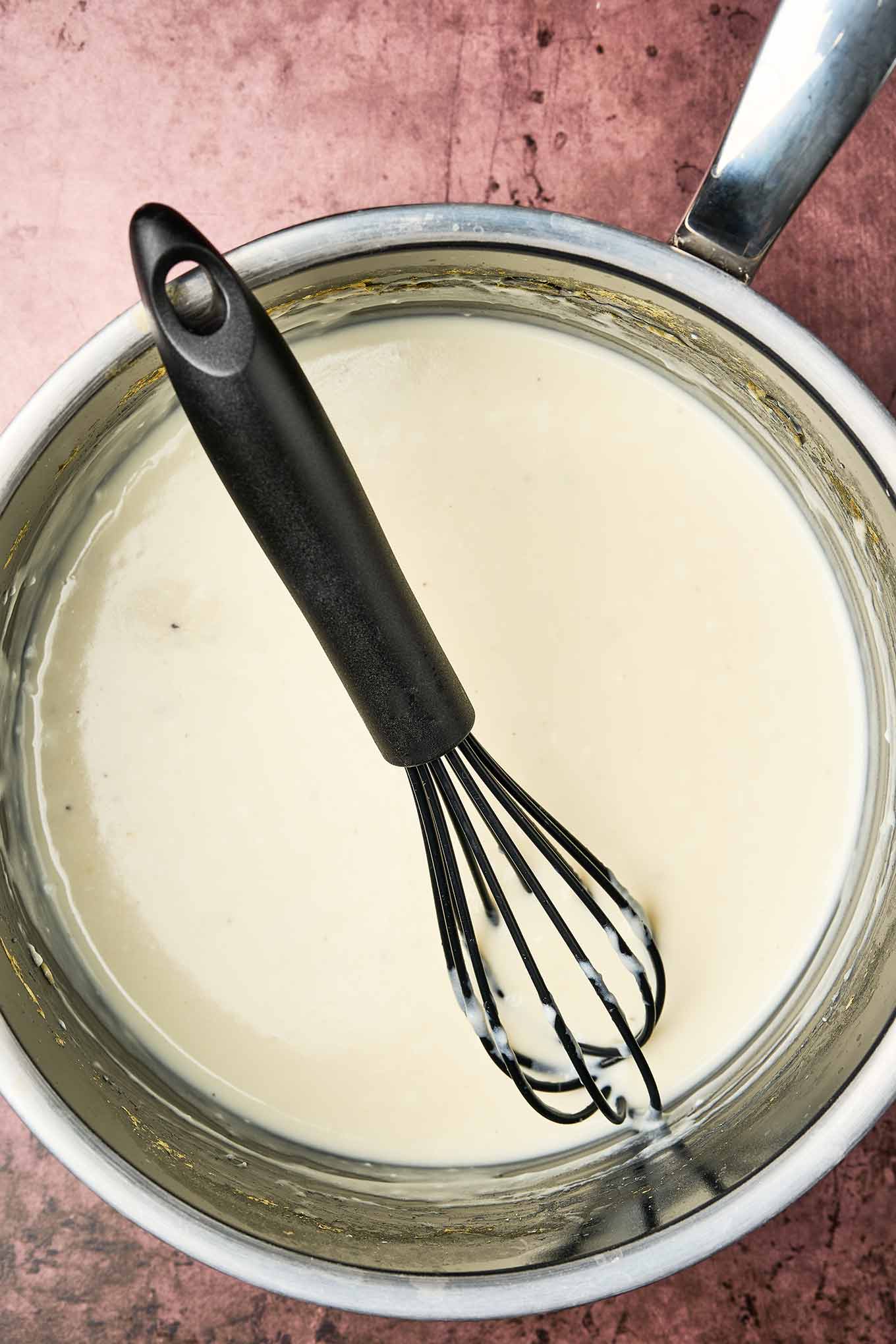 Smooth and creamy white cheese sauce in a saucepan with a black whisk, shown from above, ready to be combined with potatoes and chives and baked.