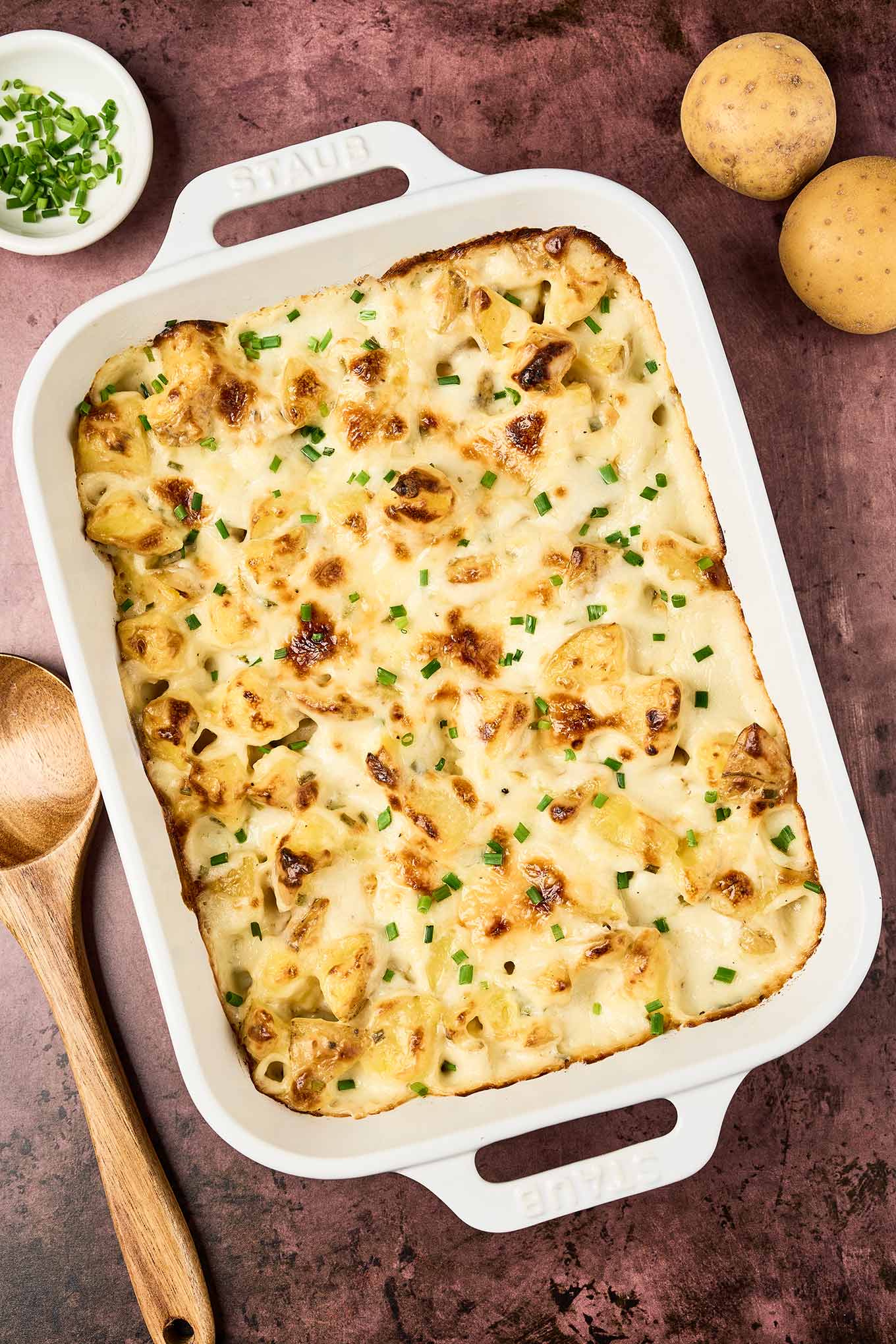 Golden-browned cottage cheese potato casserole in a white baking dish, topped with melted cheese and chopped chives, on a rustic surface with whole potatoes and a wooden spoon nearby.