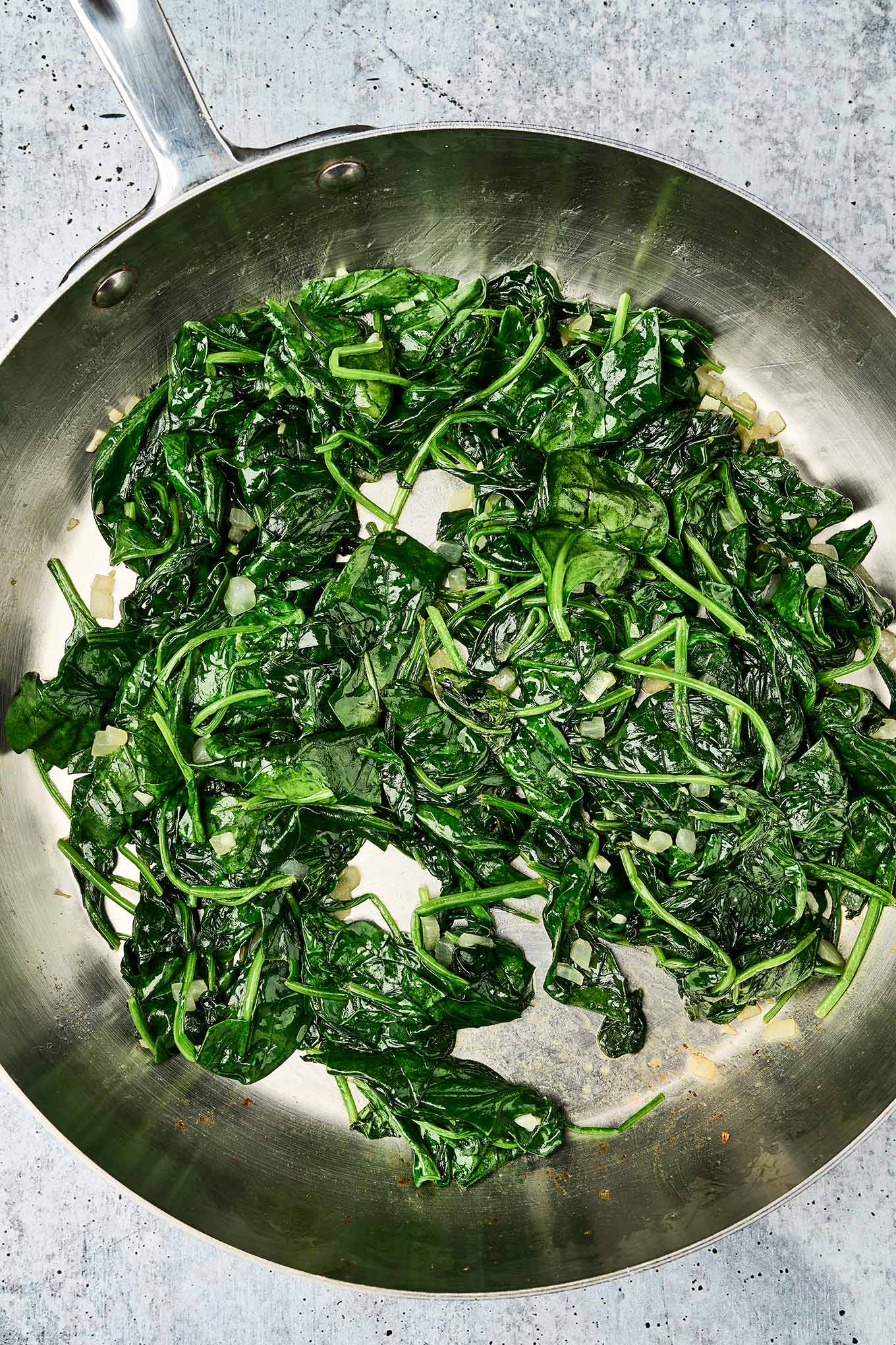 Fresh spinach saut&eacute;ed with chopped onions in a stainless steel skillet until wilted and glossy, shown on a gray countertop.