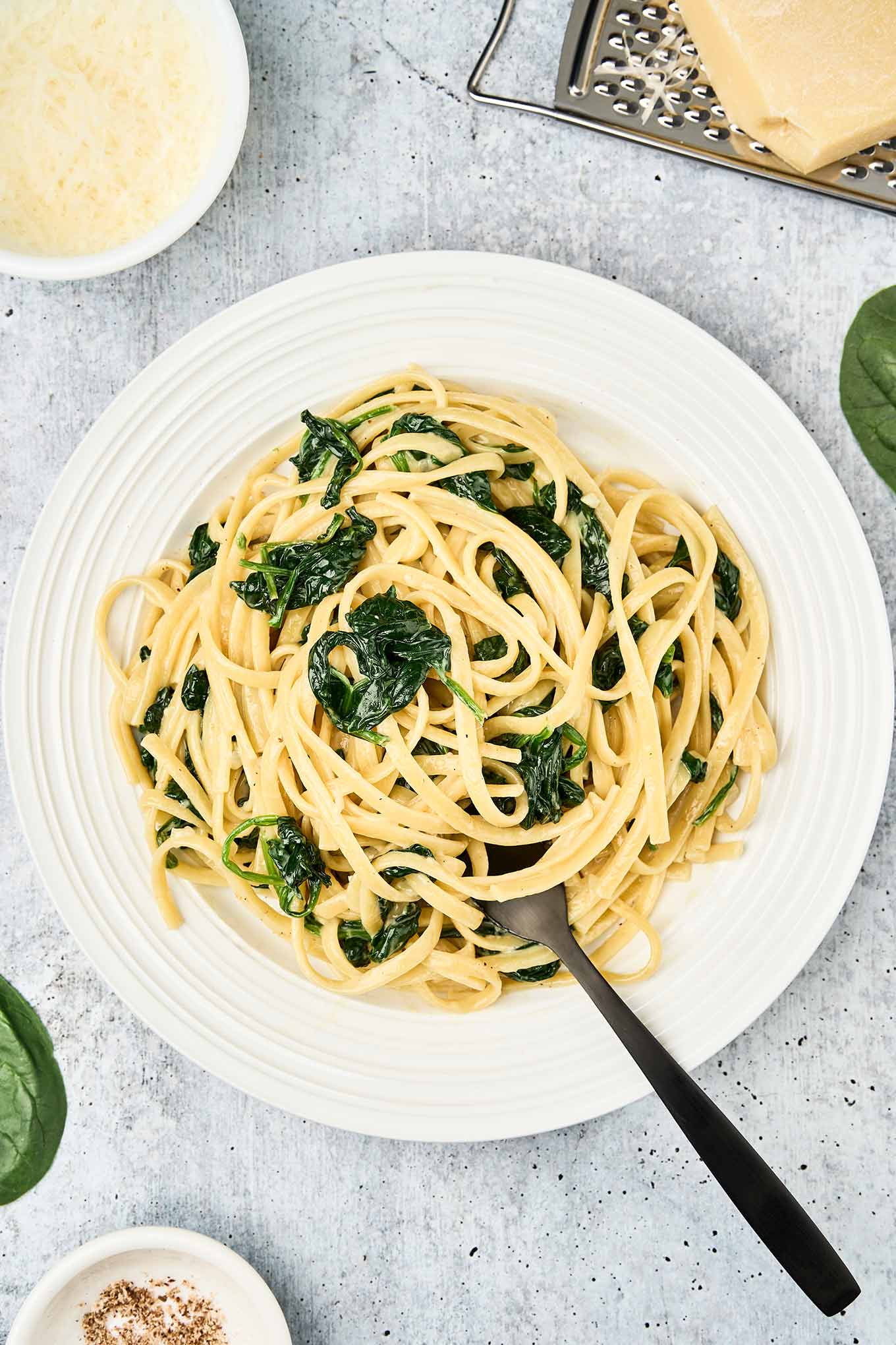 A plate of linguine tossed with saut&eacute;ed spinach and creamy mascarpone-Parmesan sauce, served with a fork on a gray surface and surrounded by cheese, nutmeg, and fresh spinach leaves.