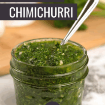 A quilted glass jam jar of chimichurri sauce made with parsley, cilantro, garlic, olive oil, wine vinegar, and seasoning, a cutting board with fresh herbs and seasonings in the background.
