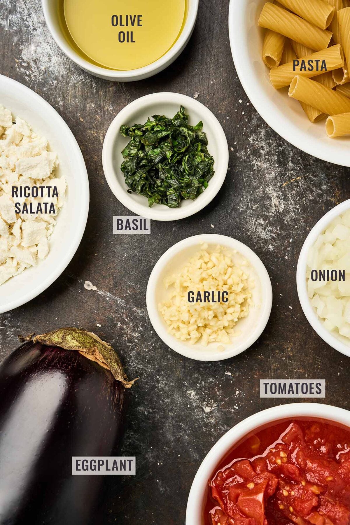 A labeled, overhead view of the ingredients for Pasta alla Norma arranged on a dark surface: a whole eggplant, chopped garlic, diced onion, canned tomatoes, fresh basil, rigatoni pasta, crumbled ricotta salata cheese, and a bowl of olive oil.