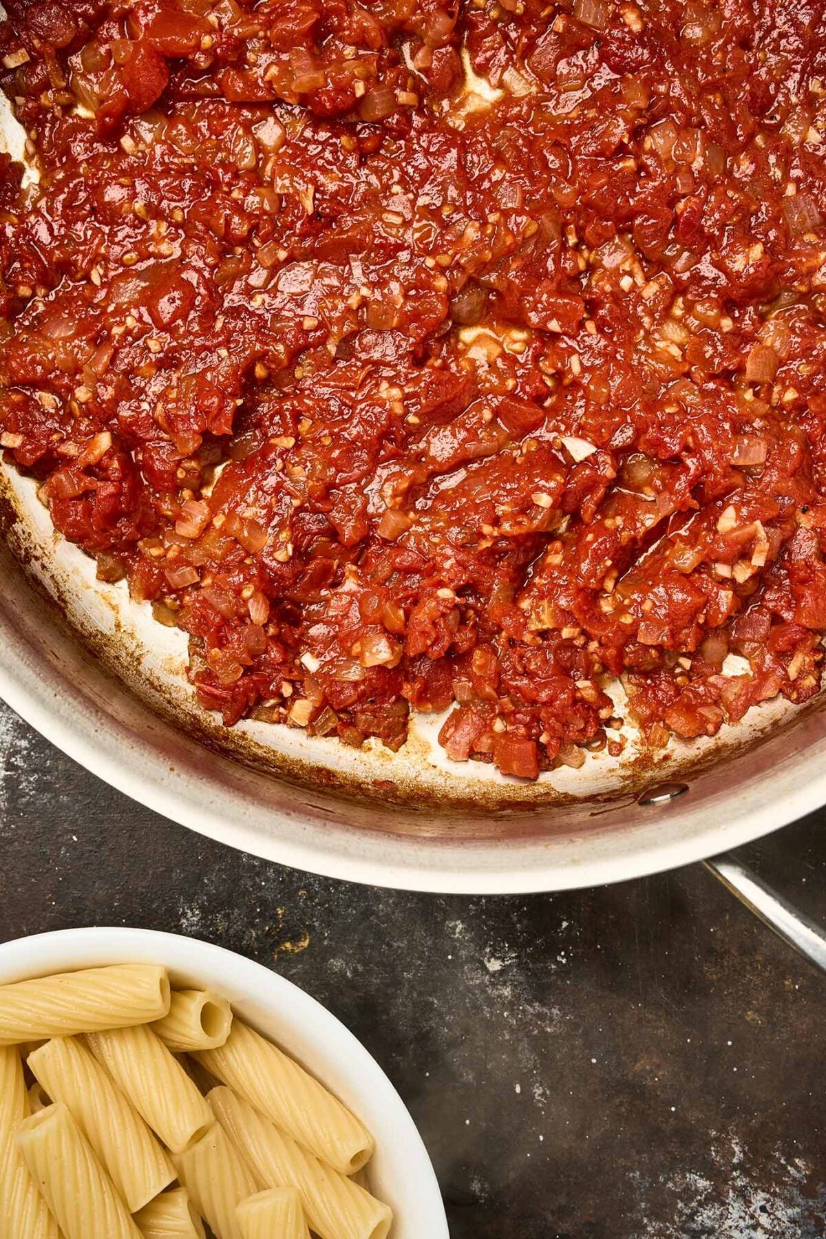 A skillet filled with tomato sauce made with diced tomatoes, garlic, and onions. Below the pan, a bowl of al dente cooked rigatoni pasta is ready to be added to the sauce.