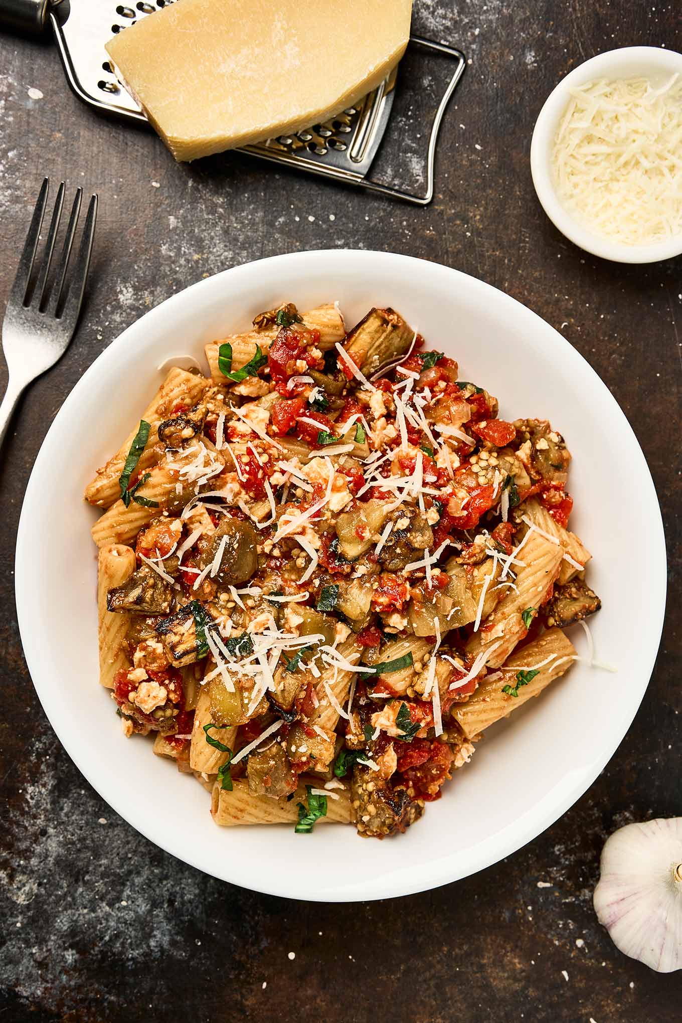 A white bowl filled with Pasta alla Norma - pasta tossed with roasted eggplant, tomato sauce, ricotta salata, and basil, topped with shredded cheese. A wedge of cheese, a grater, a fork, and a bowl of grated cheese are arranged nearby.