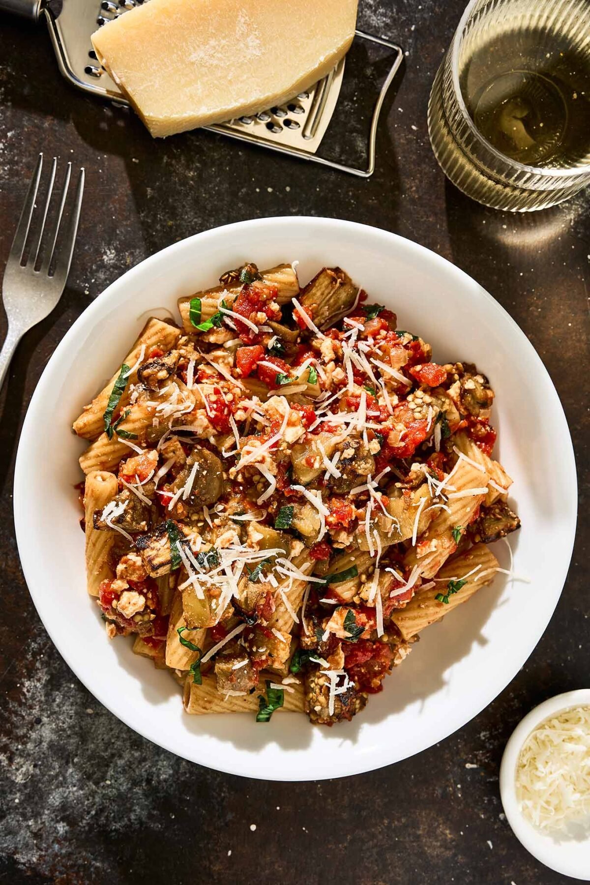 A bowl of rigatoni with roasted eggplant, tomato sauce, ricotta salata, and fresh basil topped with grated Parmesan cheese and surrounded by a wedge of cheese on a grater, a fork, a glass of white wine, and a small bowl of extra grated cheese.