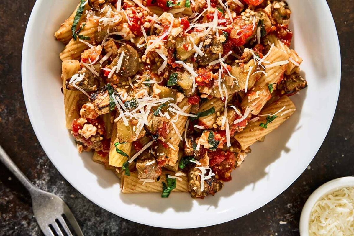 A close-up of Pasta alla Norma in a white bowl, featuring rigatoni tossed with roasted eggplant, chunky tomato sauce, fresh basil, and ricotta salata. A fork and small dish of grated cheese sit nearby on a dark surface.