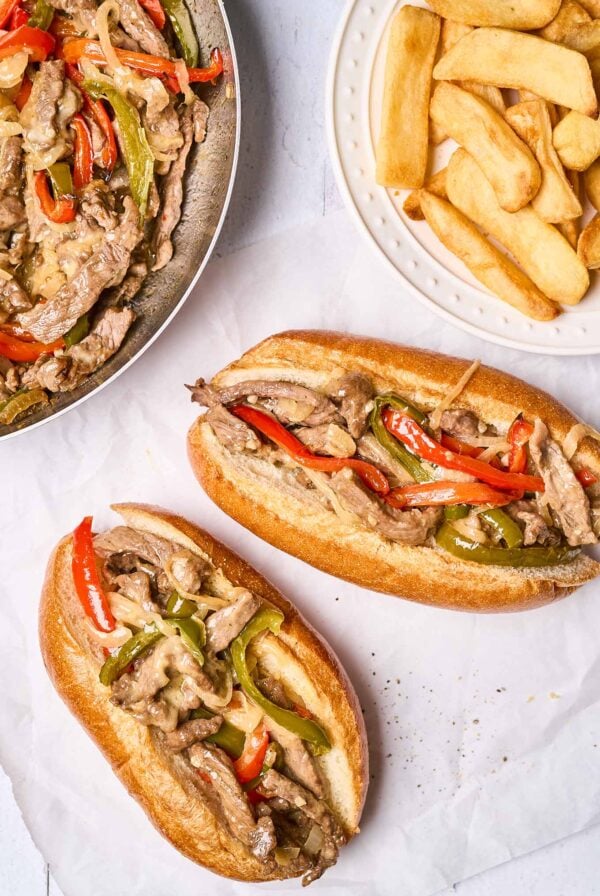 Two Philly cheesesteak sandwiches with peppers, onions, and provolone cheese on toasted sub rolls, a plate of steak fries and the frying pan with more steak, peppers, and onions in the background.