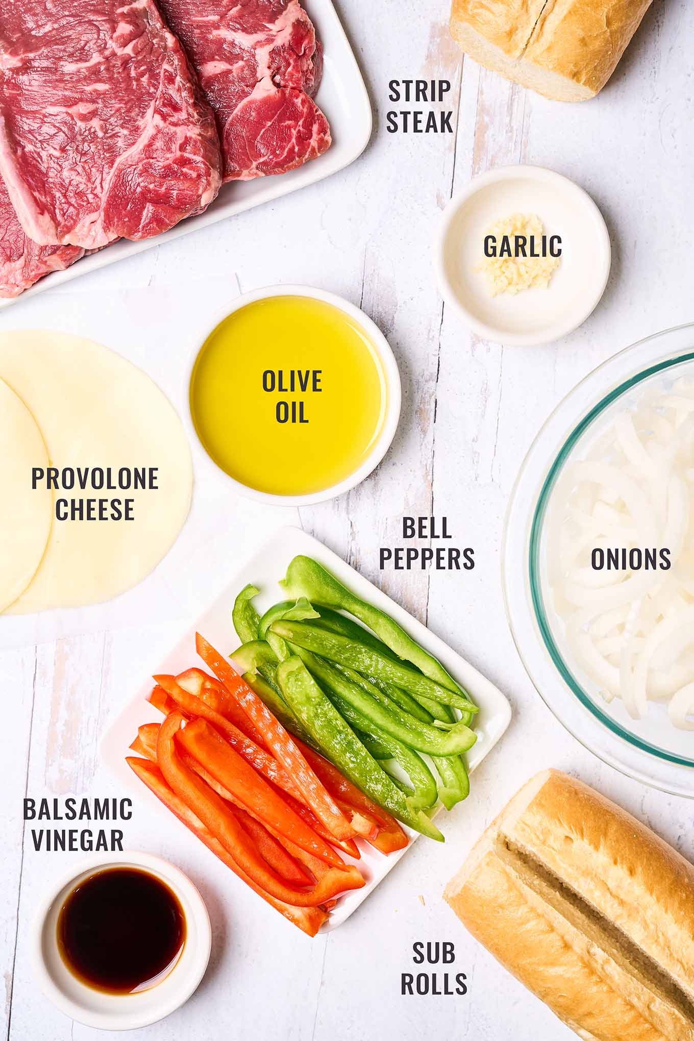 A labeled, overhead view of the ingredients needed to make philly cheesesteak sandwiches at home; strip steak, sub rolls, provolone cheese, olive oil, garlic, onions, red and green bell peppers, and balsamic vinegar.