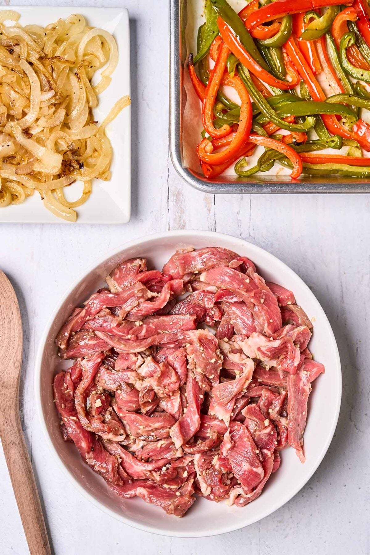 A parchment-lined sheet pan with strips of roasted red and green bell pepper, a plate filled with sautéed onions, and a bowl filled with marinated strips of steak, ready for pan-searing.
