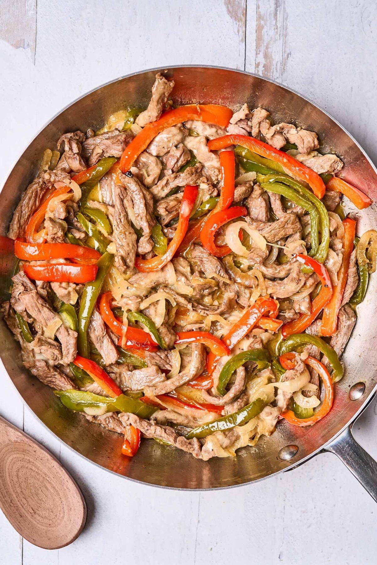 A frying pan filled with strips of pan-seared steak, roasted bell pepper, sautéed onions, and melted provolone cheese - ready to be spooned into sub rolls, a wooden spoon in the background.
