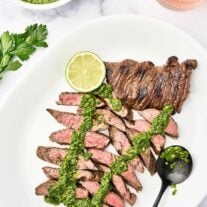 A white serving platter with sliced, medium-rare, grilled skirt steak, topped with chimichurri sauce, garnished with a lime slice, a bowl of chimichurri and glass of rosé in the background.