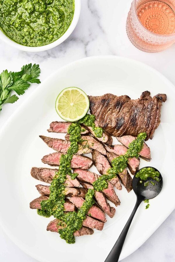 A white serving platter with sliced, medium-rare, grilled skirt steak, topped with chimichurri sauce, garnished with a lime slice, a bowl of chimichurri and glass of rosé in the background.