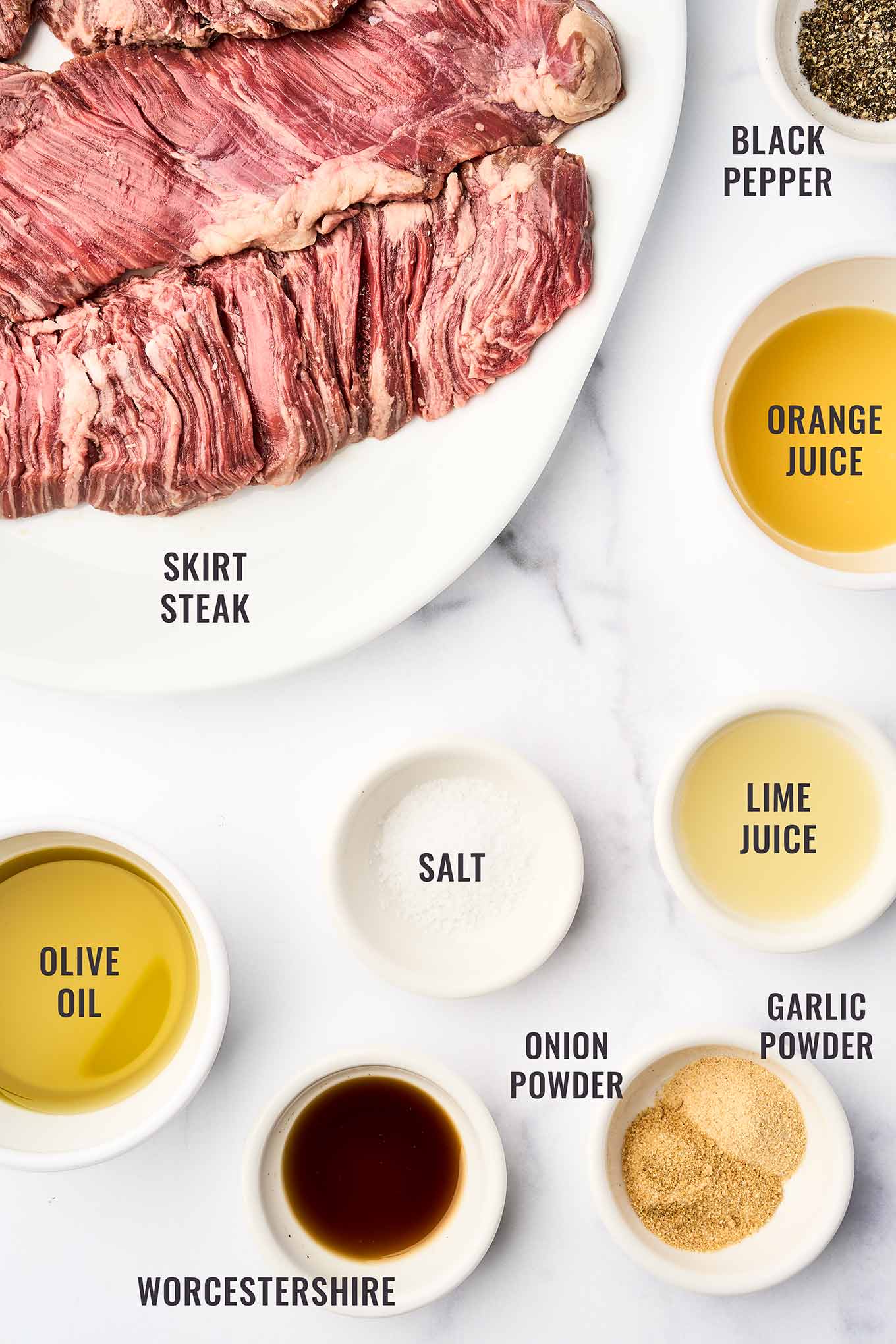 A labeled, overhead view of the marinade ingredients for grilled skirt steak ingredients including skirt steak, orange juice, lime juice, olive oil, Worcestershire sauce, garlic powder, onion powder, salt, and black pepper.