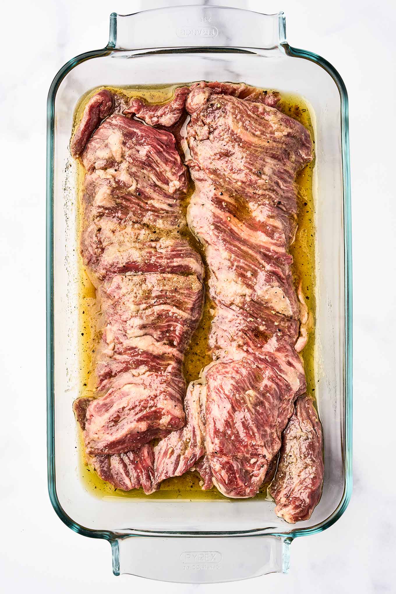 Raw skirt steak marinating in a glass dish with olive oil, orange and lime juice, and seasonings - the first step for making grilled skirt steak with chimichurri sauce.