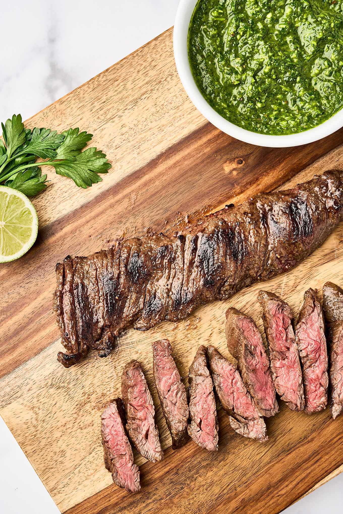 Grilled skirt steak partially sliced on a wooden cutting board, ready to be topped with chimichurri sauce from the bowl beside it.