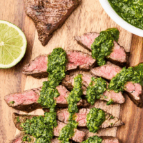 Sliced grilled skirt steak drizzled with vibrant green chimichurri sauce on a wooden cutting board, served with fresh lime.