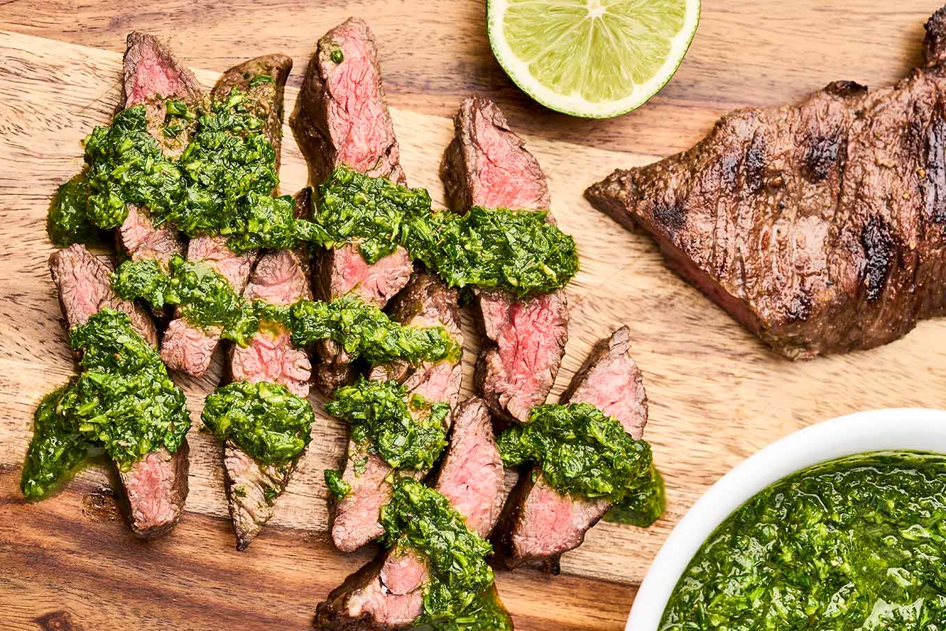 Sliced grilled skirt steak drizzled with vibrant green chimichurri sauce on a wooden cutting board, served with fresh lime.