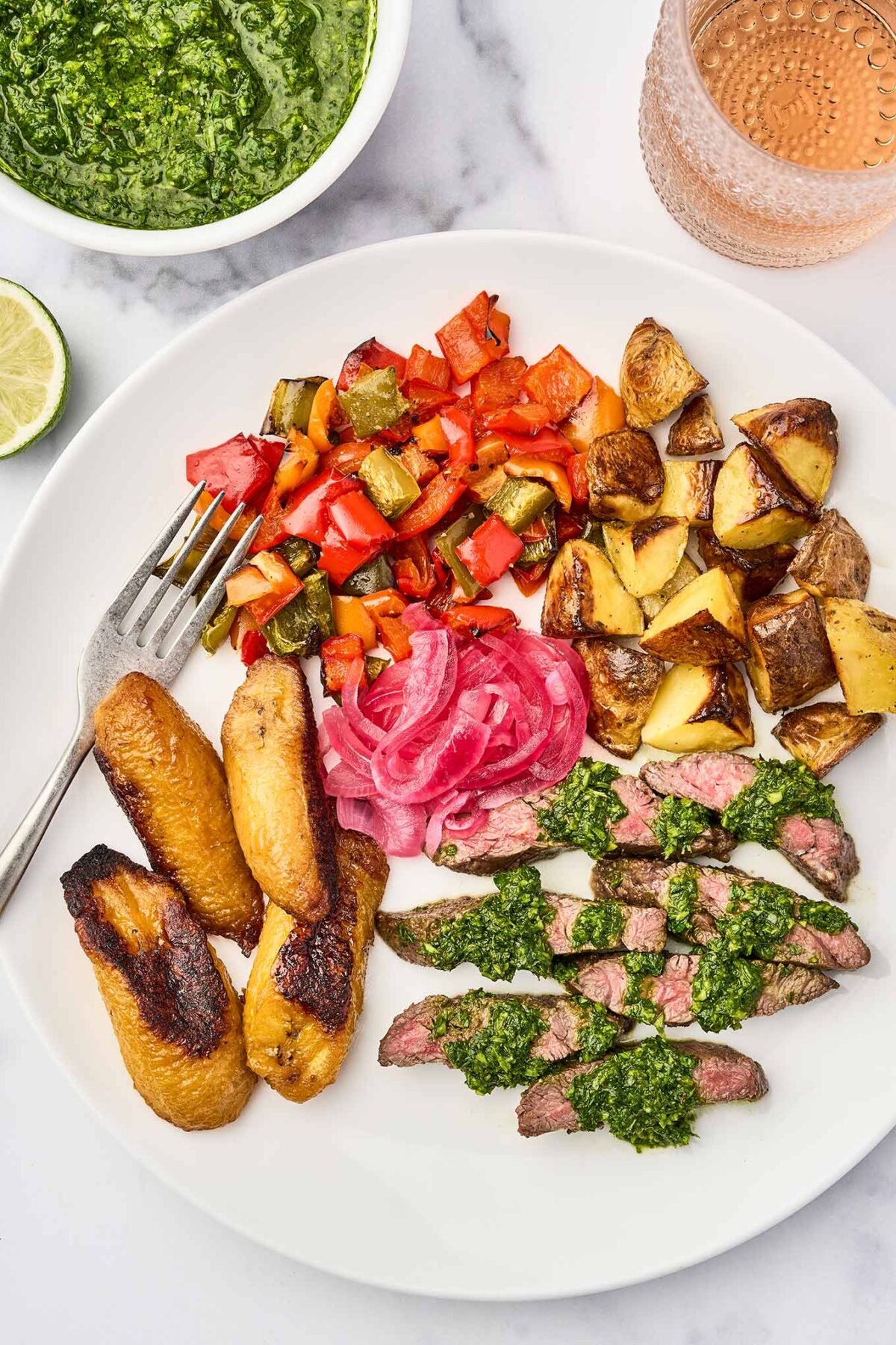 A plate of grilled skirt steak with chimichurri sauce, served with roasted sweet plantains, pickled red onions, roasted potatoes, and sautéed bell peppers.