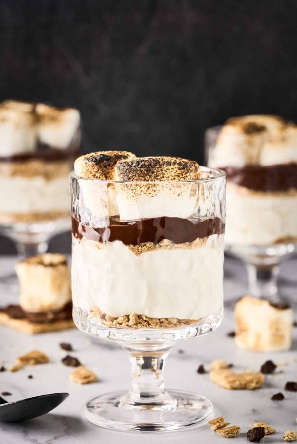A s'mores cheesecake parfait in a clear glass surrounded by more parfaits, extra toasted marshmallows, bits of graham cracker crumbs, and chunks of chocolate.