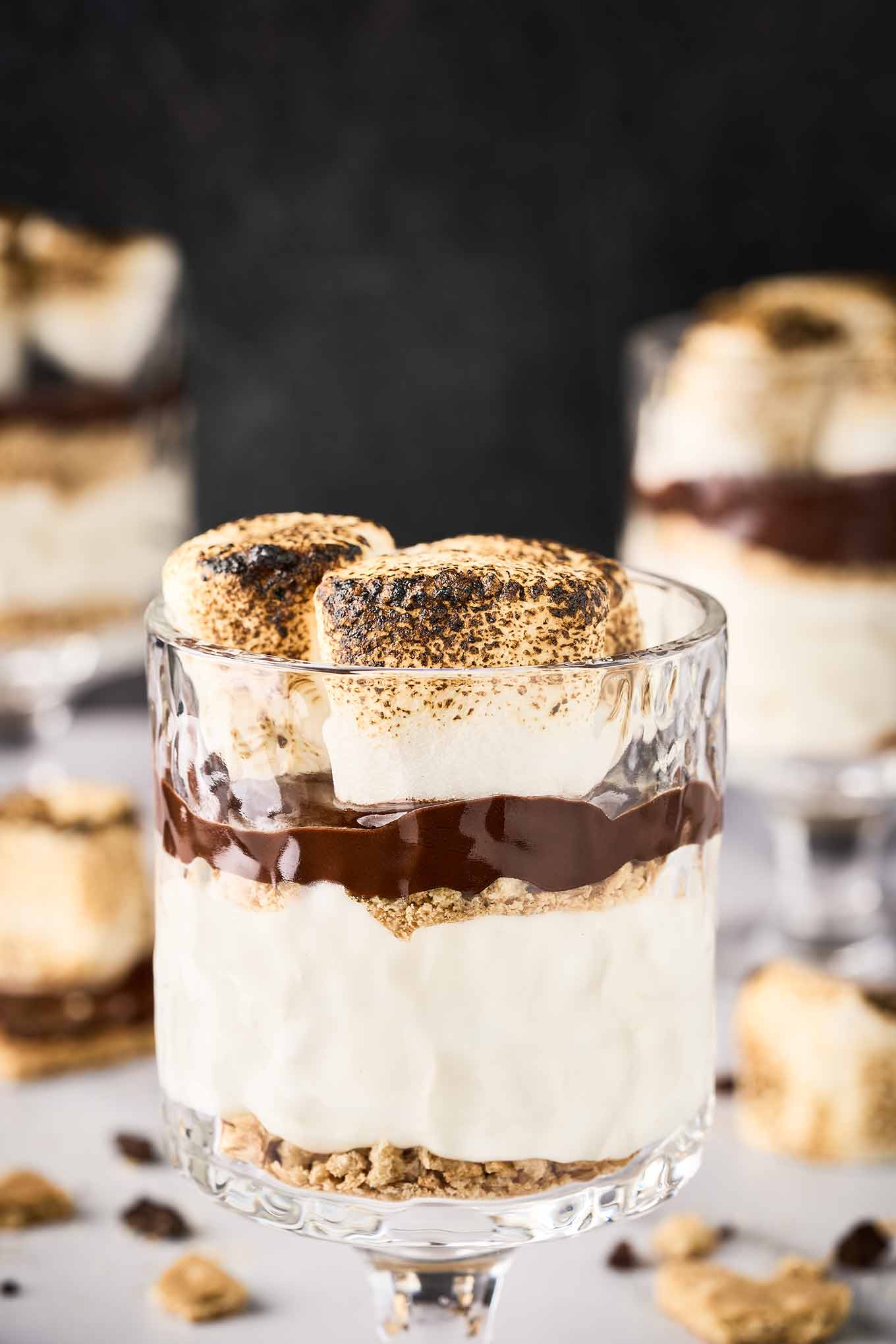 A footed, clear dessert glass filled with s'mores cheesecake parfait with layers of cheesecake filling, graham cracker crumbs, chocolate ganache, and toasted marshmallows.
