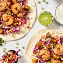 Spicy shrimp tacos with sweet plantains, tropical slaw, and lime crema on flour tortillas, garnished with cilantro and scallions.