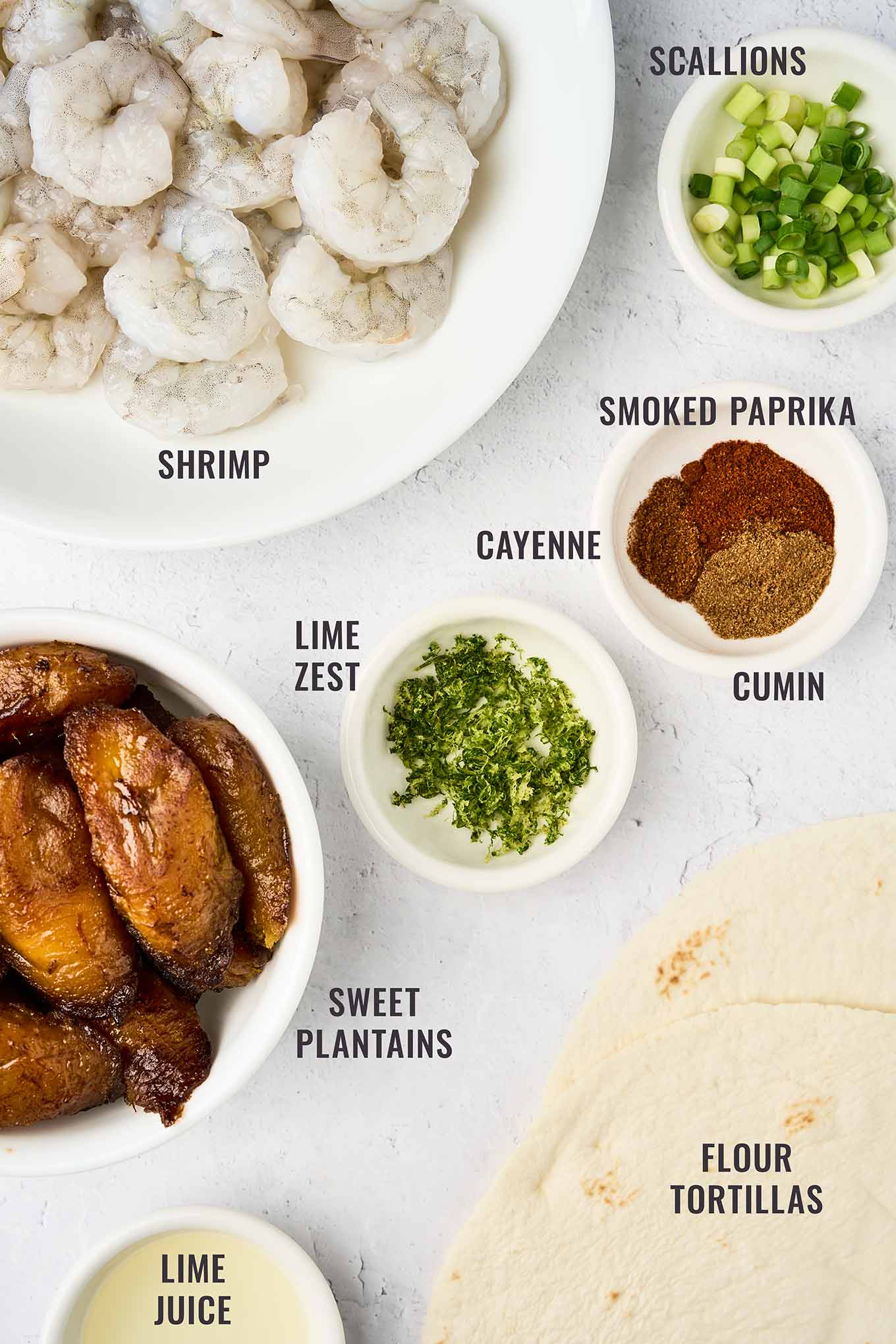 A labeled, overhead view of the ingredients for spicy shrimp tacos with sweet plantains and tropical slaw, including shrimp, scallions, lime zest and juice, sweet plantains, flour tortillas, and assorted spices.