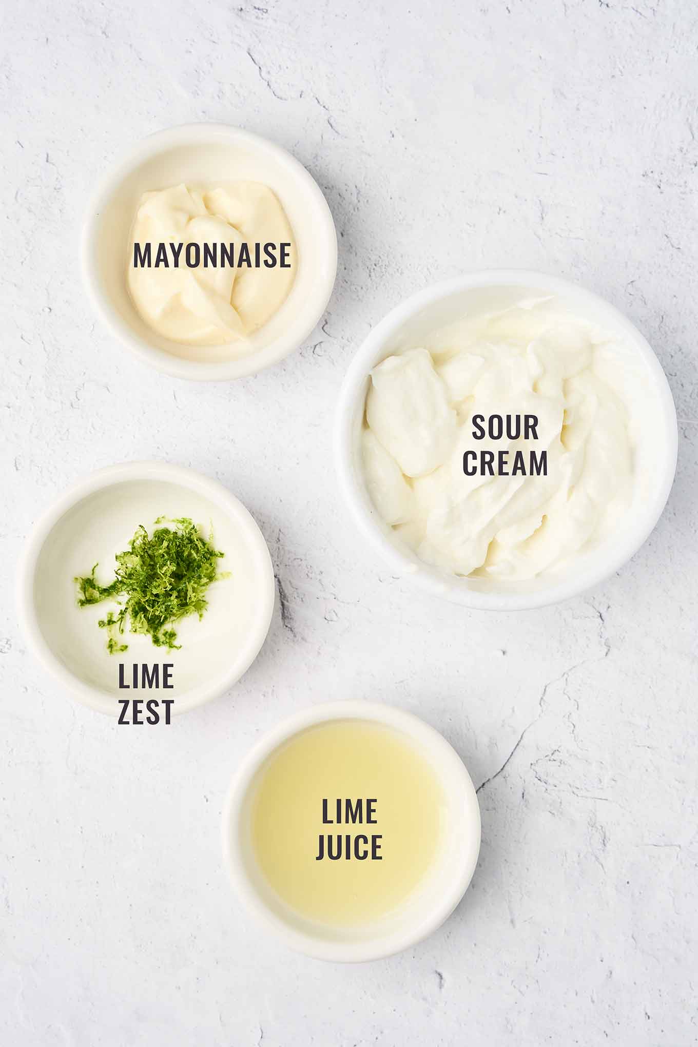 A labeled, overhead view of the ingredients for lime crema topping for shrimp tacos, including mayonnaise, sour cream, lime zest, and lime juice - all in small white bowls.