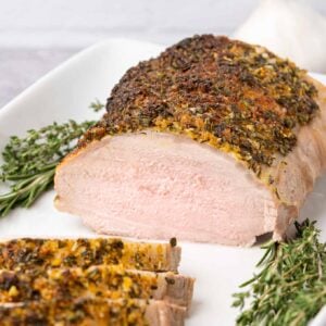 Garlic and herb crusted boneless pork roast sliced on a white plate with fresh rosemary and thyme garnish.