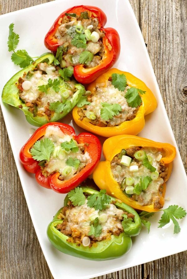 A white serving platter with red, green, and yellow bell pepper halves filled with a Mexican-spiced mix of ground beef, tomatoes, and rice, topped with melted cheese, and garnished with fresh cilantro.