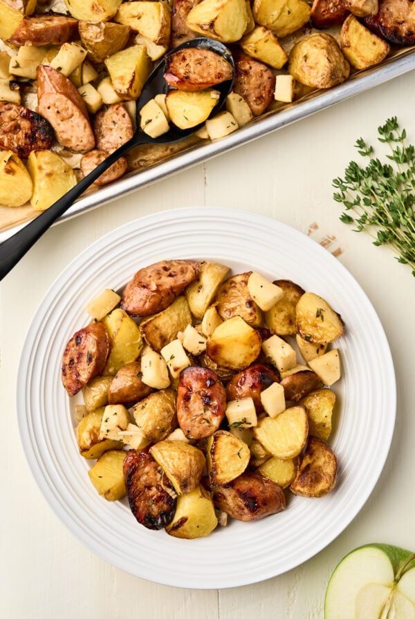 Roasted chicken sausage with apples and potatoes served on a white plate with fresh thyme and a sliced apple on the side.