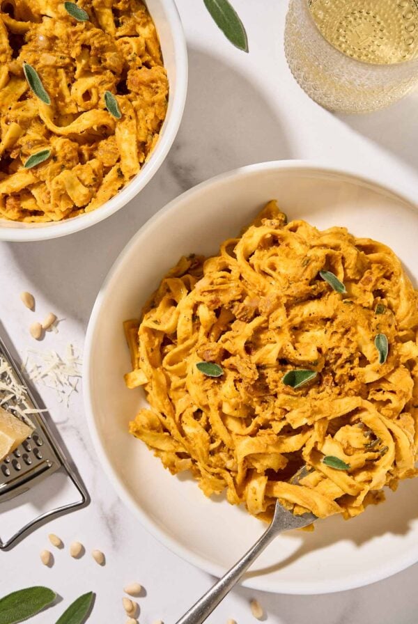 Two bowls of creamy pumpkin pasta with pancetta and sage served in white bowls, a glass of white wine and cheese grater in the background.