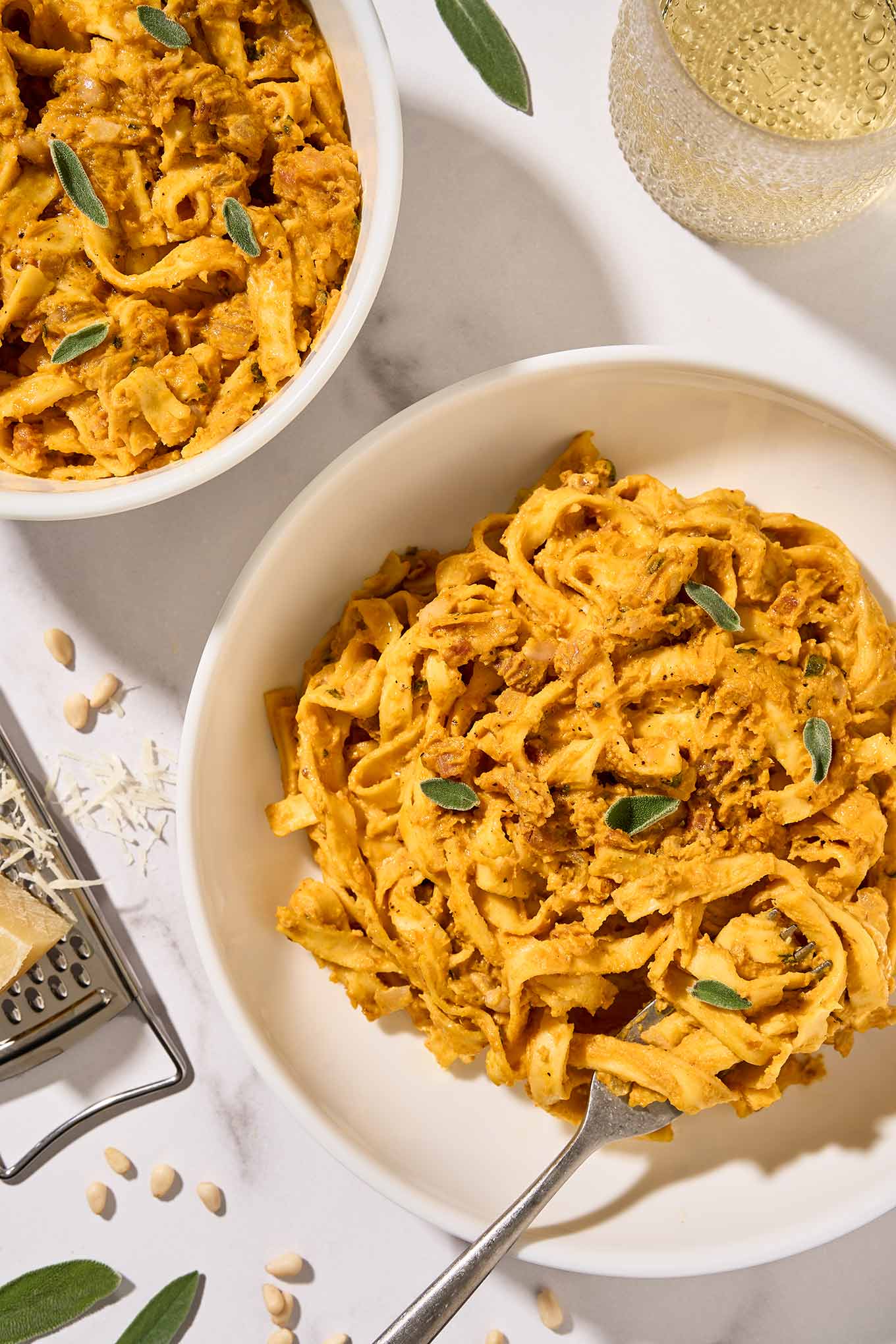 Two bowls of creamy pumpkin pasta with pancetta and sage served in white bowls, a glass of white wine and cheese grater in the background.