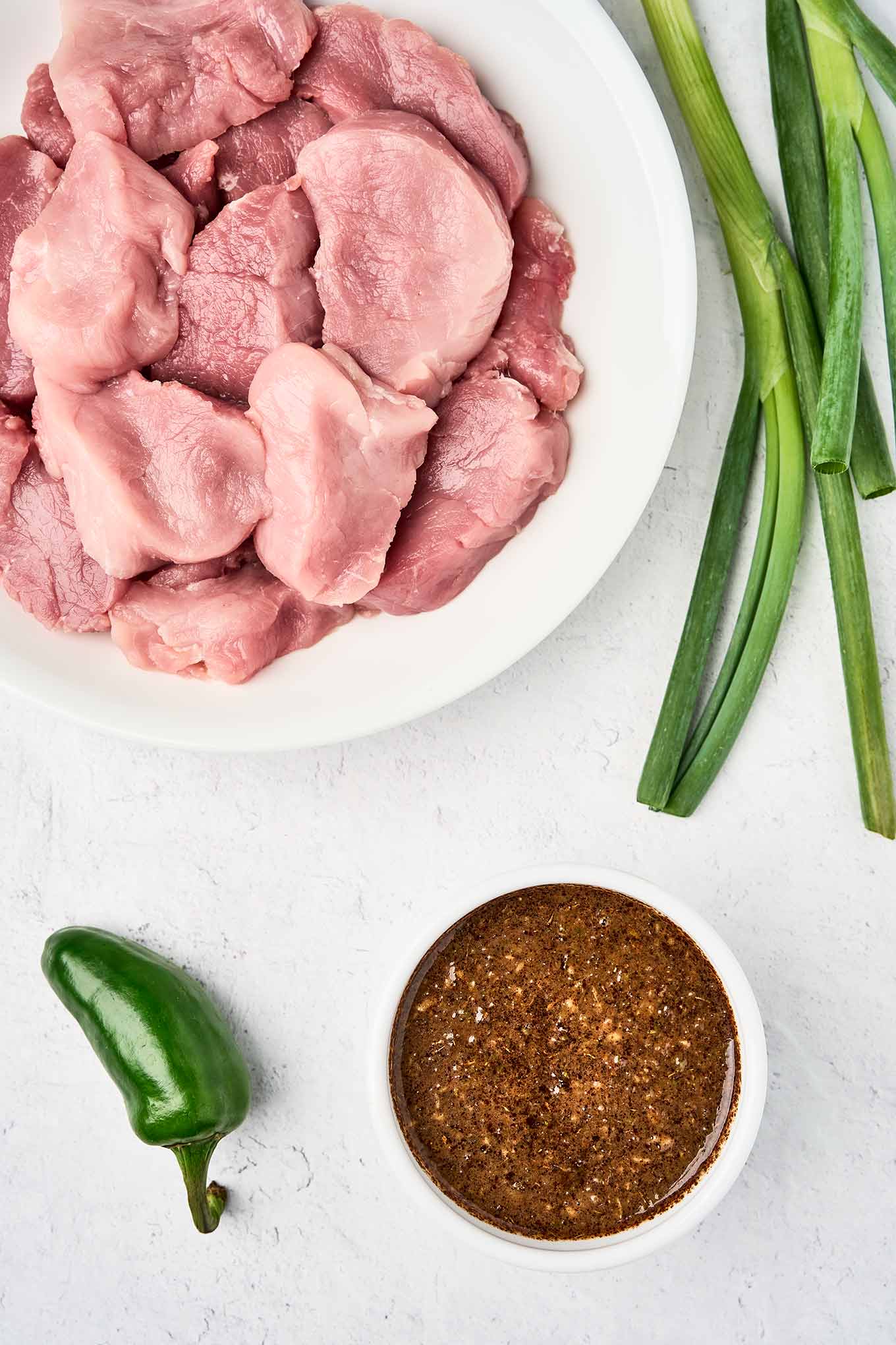 Whole scallions and a jalapeño pepper ready to be pureed with other jerk marinade ingredients, pork tenderloin medallions on a white plate in the upper left.