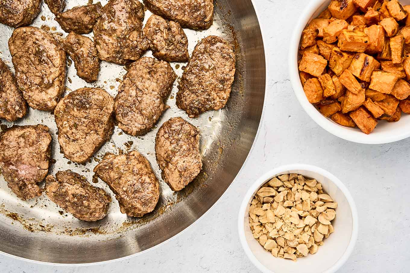 Seared jerk pork tenderloin medallions in a skillet, with bowls of roasted sweet potato cubes and chopped toasted peanuts alongside.