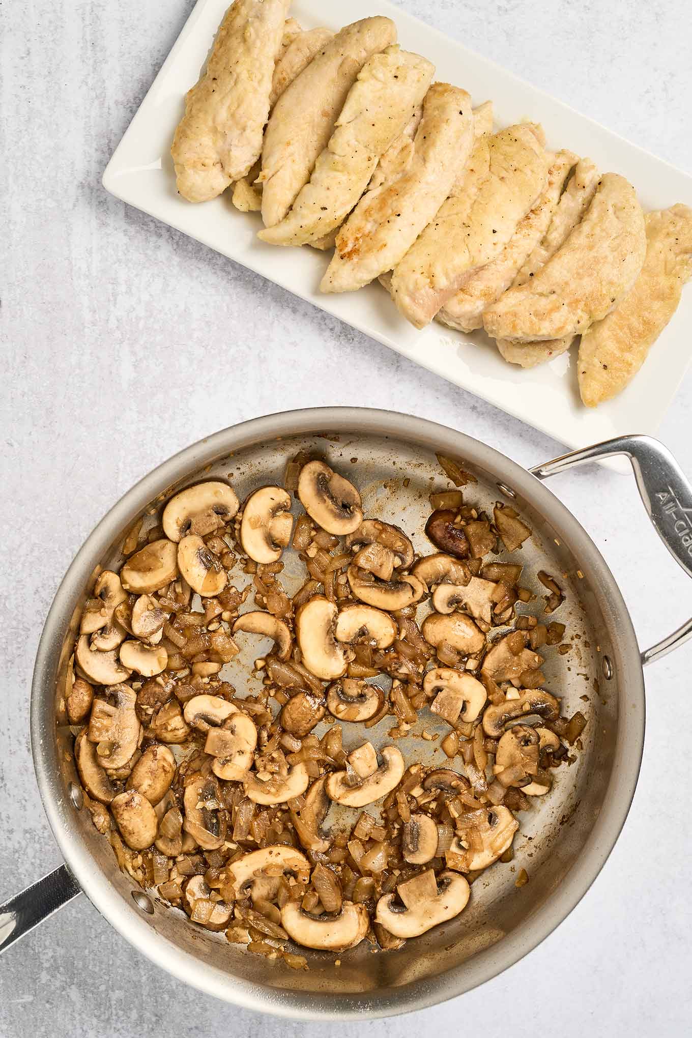 Partially cooked chicken tenderloins on a plate beside a skillet of mushrooms, onion, and garlic for mushroom chicken.