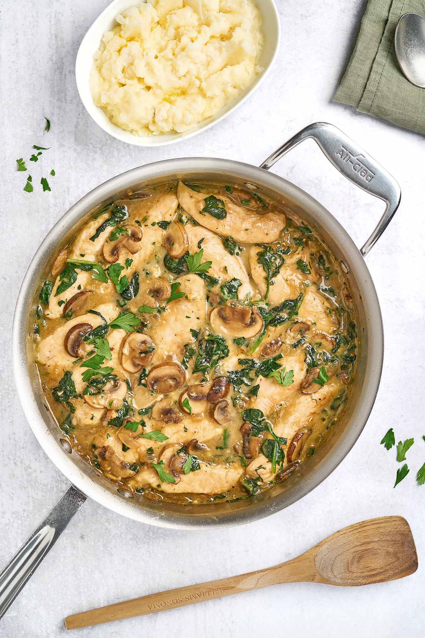 A sauté pan of chicken tenderloins simmered in a savory sauce with sautéed mushrooms, wilted spinach, and a garnish of fresh parsley, served alongside a bowl of mashed potatoes.