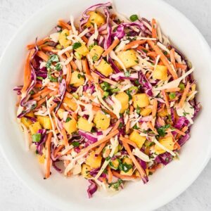 A white bowl of pineapple slaw made with red and green cabbage, shredded carrots, chunks of fresh pineapple, scallions, cilantro, and a tangy dressing.