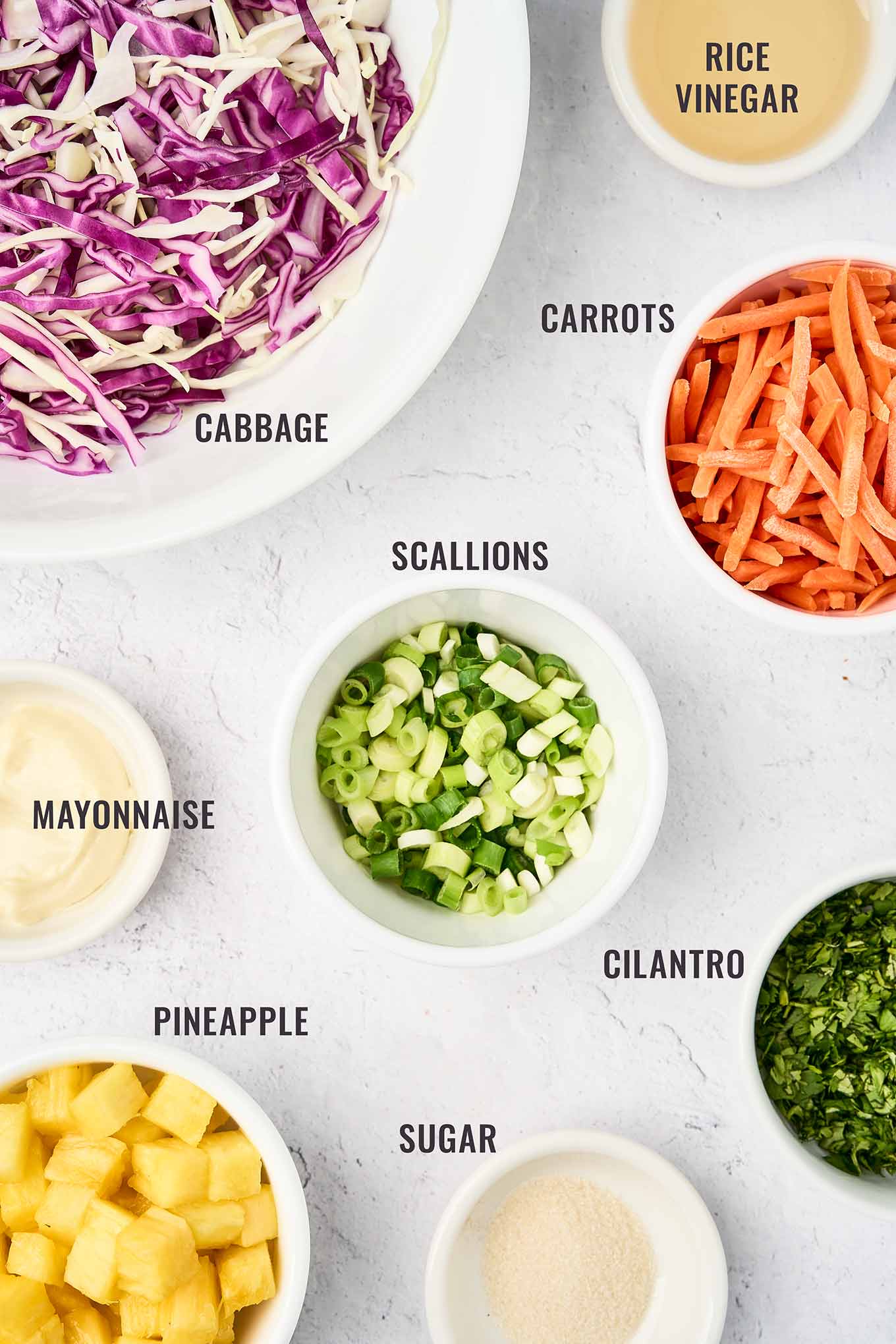 A labeled, overhead view of the ingredients for pineapple slaw; shredded cabbage, shredded carrots, scallions, cilantro, pineapple cubes, sugar, mayo, and rice vinegar.