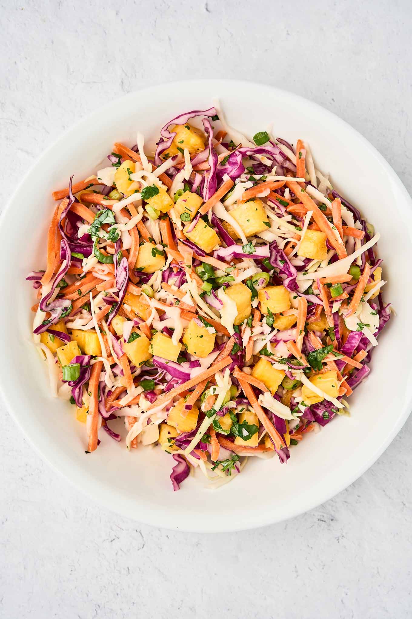 A white bowl of pineapple slaw made with red and green cabbage, shredded carrots, chunks of fresh pineapple, scallions, cilantro, and a tangy dressing.
