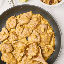A skillet of pan seared pork tenderloin medallions and chunks of sautéed apple in a mild, creamy curry sauce, being stirred with a wooden spoon. A bowl of pearl couscous for serving in the background.