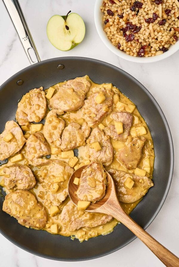 A skillet of pan seared pork tenderloin medallions and chunks of sautéed apple in a mild, creamy curry sauce, being stirred with a wooden spoon. A bowl of pearl couscous for serving in the background.