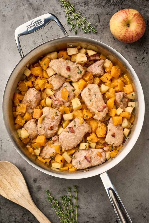 Pork medallions with butternut squash and apples simmered in a maple-thyme sauce with bacon, served in a stainless-steel skillet.