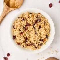 A bowl of toasted pearl couscous with dried cranberries and slivered almonds surrounded by a wooden spoon, scattered cranberries, and a wooden measuring cup with uncooked Israeli couscous.