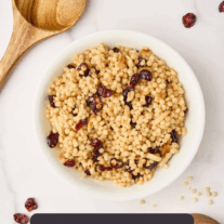 A bowl of toasted pearl couscous with dried cranberries and slivered almonds surrounded by a wooden spoon, scattered cranberries, and a wooden measuring cup with uncooked Israeli couscous.